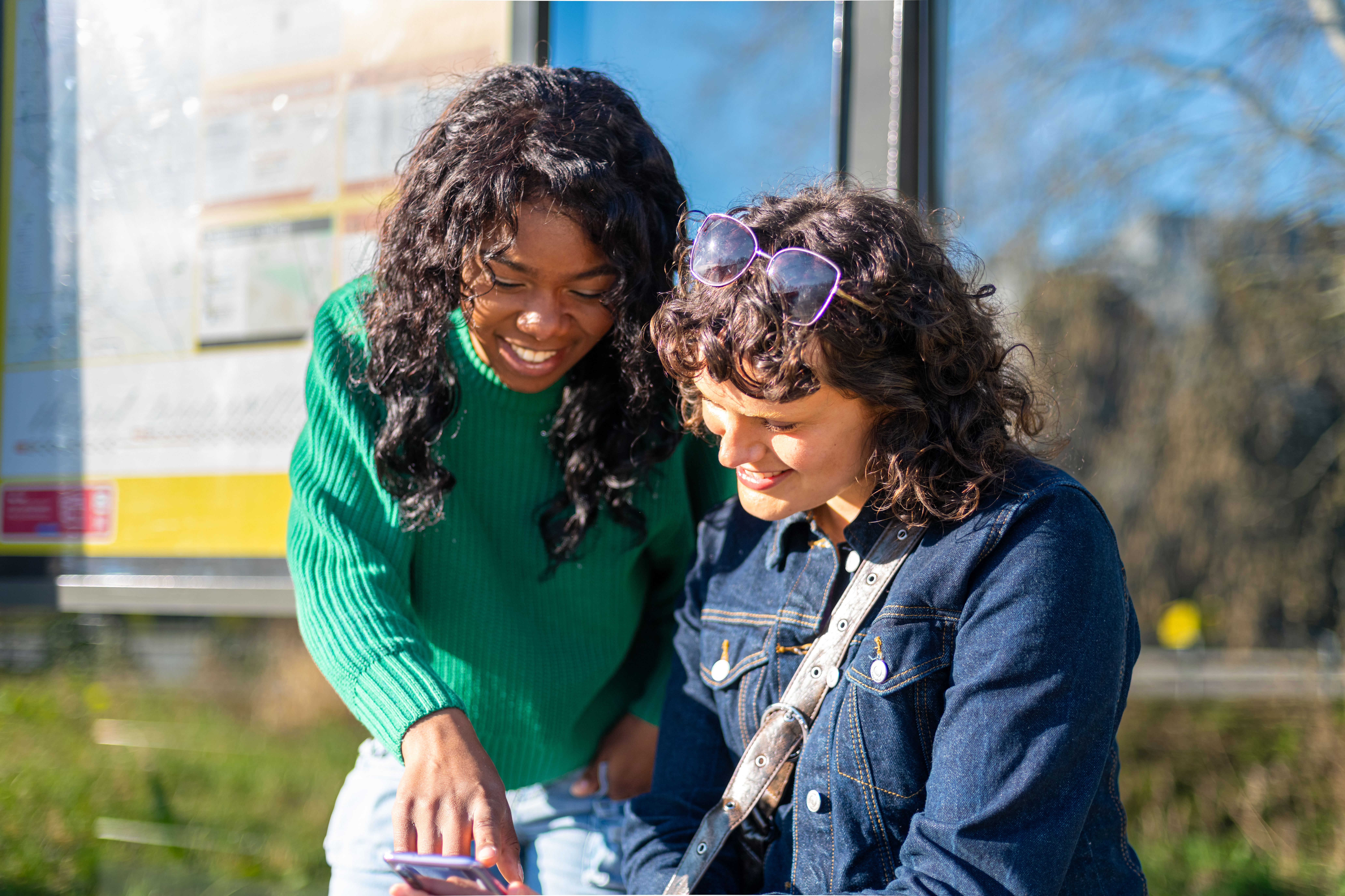 Twee jonge vrouwen die bij een bushalte staan en kijken op de telefoon van één van de twee.