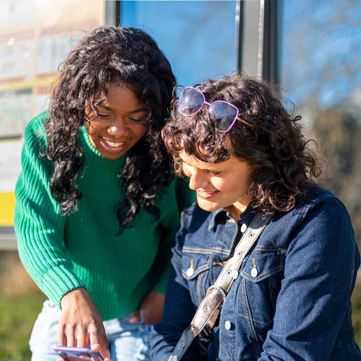 Two young women standing at a bus stop looking at one of their phones.