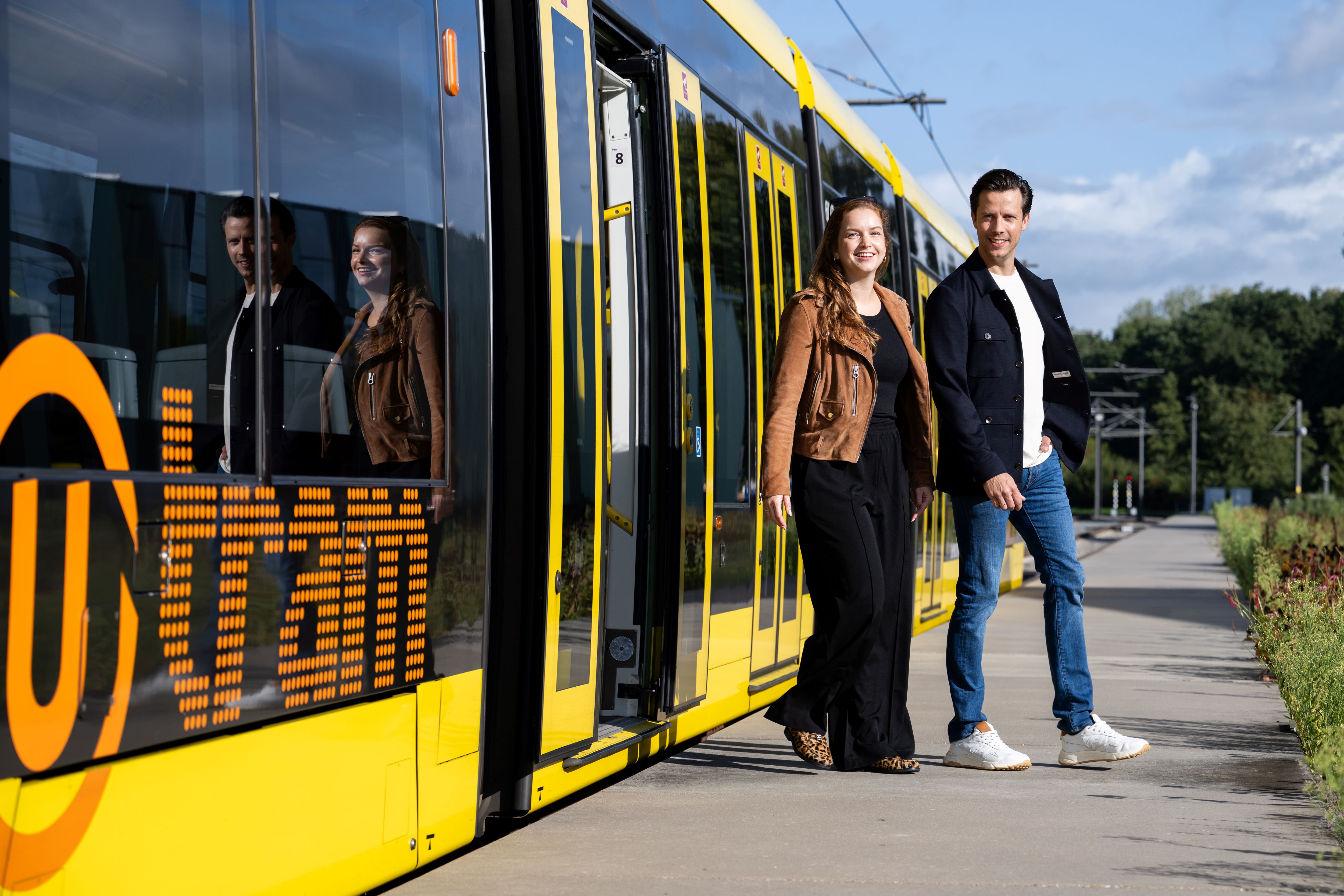 Twee personen die uit een gele tram stappen op een perron. De tram heeft het oranje UOV logo.