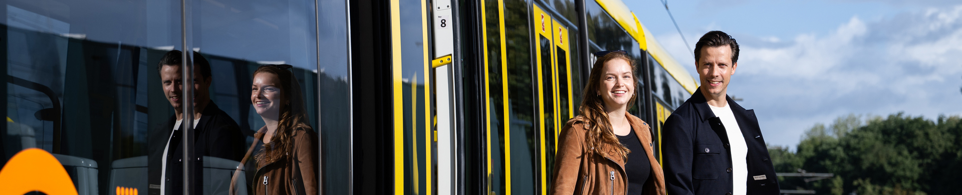 Two people stepping off a yellow tram onto a platform. The tram has the orange UOV logo.