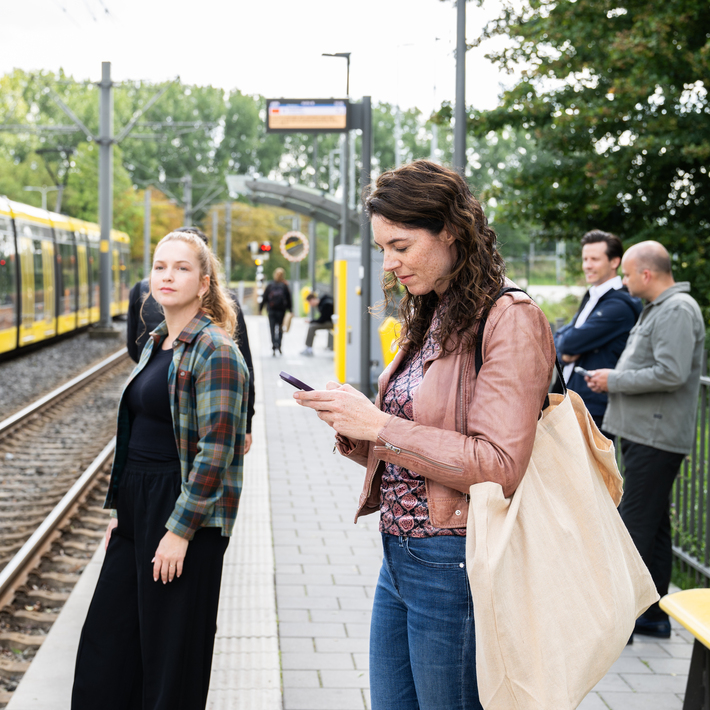 Passengers at a tram stop. One woman is looking at her phone, the other is looking towards the camera.