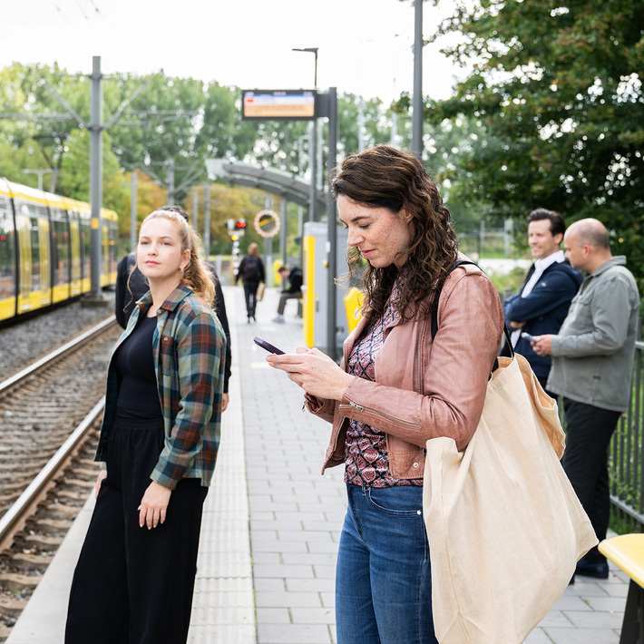 Reizigers bij tramhalte. Één vrouw kijkt op haar telefoon, de andere richting de camera.