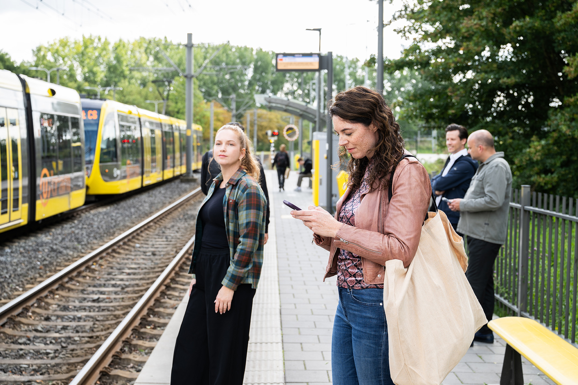 Reizigers bij tramhalte. Één vrouw kijkt op haar telefoon, de andere richting de camera.