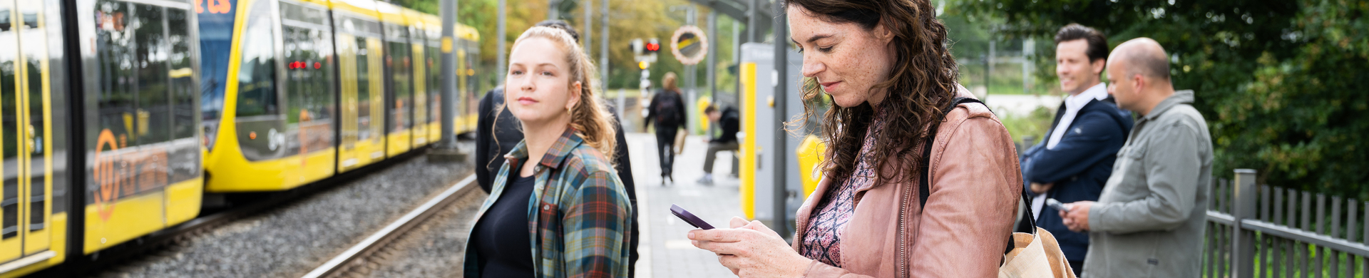 Reizigers bij tramhalte. Één vrouw kijkt op haar telefoon, de andere richting de camera.